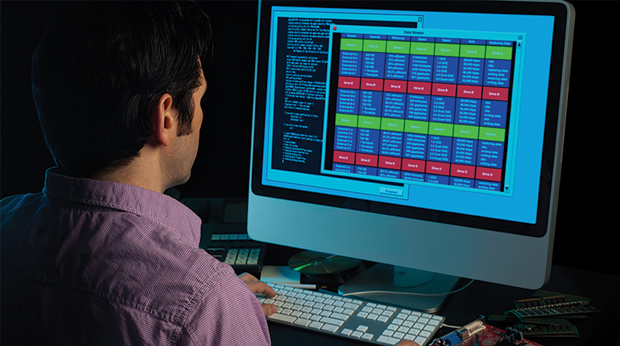 A person sits in front of a desktop computer. The screen has multiple color charts and coding on the left side of the screen.